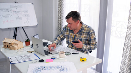 The designer is sitting at a desk with a laptop, building drawings and a mobile phone. He is focused on working on the project