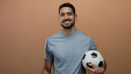 Young man smiling while holding soccer ball in front of plain brown background highlighting casual sporty lifestyle