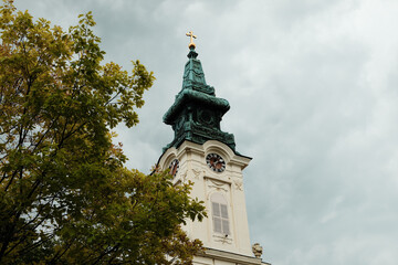 Church tower with clock and red-tiled roof partially covered by tree branches in Sombor, Serbia....