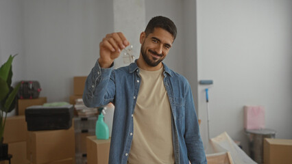 Young man holding house keys smiling in a new home interior surrounded by boxes indicating moving...