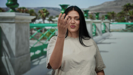 Woman with long dark hair blows a kiss to the camera while standing on a city street with green railings and palm trees in the sunny background.