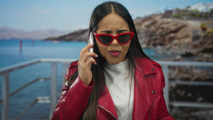 Woman using a phone displays an angry look against a beach backdrop featuring a young latin...