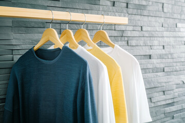 Obraz premium Blue, white and yellow t-shirts hanging on wooden hangers in a modern wardrobe with a gray stone wall