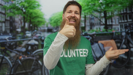 Smiling bearded man wearing volunteer shirt shows thumbs up gesture on bike lined city street;...