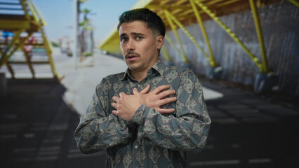Young hispanic man outdoors city street looking scared with hands on chest under bright sunlight...