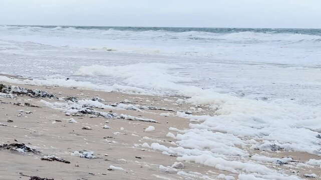 Rough ocean waves crash on Christies Beach, South Australia, covered in dense white foam from a toxic algal bloom. This environmental impact affects marine life and human health.