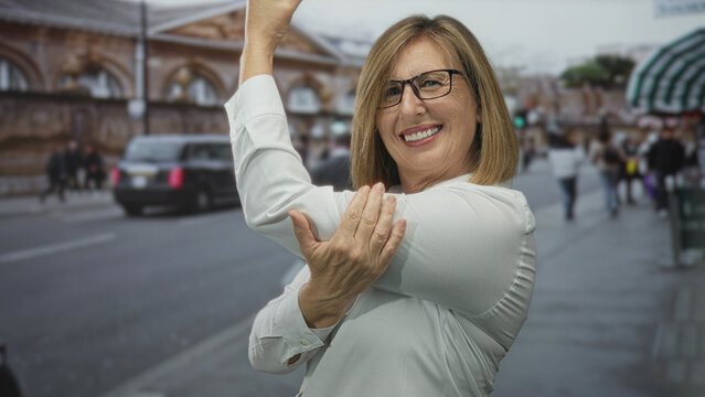 Middle aged caucasian woman in white shirt flexes arm on city street past blurred buildings and people; confidence empowerment.