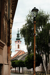 Church tower with a green dome and clock peeking through buildings in the old town of Sombor,...