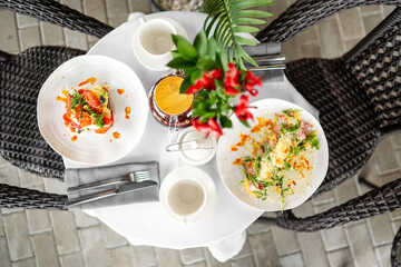 Top view of a cozy breakfast table setup with colorful dishes, cups, teapot, and a flower vase on a white tablecloth, surrounded by wicker chairs.
