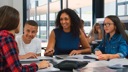Female High School Teacher Sitting At Desk Helping Students