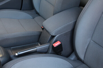 Armrest of a modern car located between the front seats. Detailed view of cars center console: armrest with stitching, seat belt, handbrake with leather handle and seats with gray fabric upholstery.