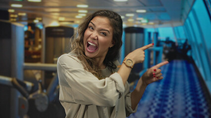 Hispanic woman in a gym enthusiastically pointing with bright expression, surrounded by fitness...