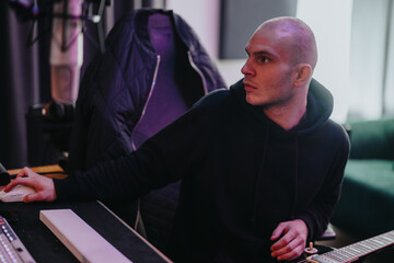 A man in a black hoodie works diligently at a mixing console in a recording studio. He concentrates...