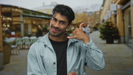 Hispanic man making phone gesture on outdoor restaurant terrace with tables and chairs visible;...