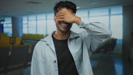 Young hispanic man covers his eyes with hand in airport terminal while smiling broadly; playfulness.