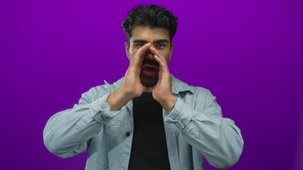 Man shouting with hands cupping mouth on a purple studio set, wearing a light denim shirt and...