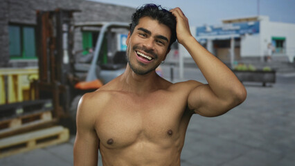 Shirtless man with muscular chest smiles and scratches head next to a forklift in front of a building on a sunny day; joy.