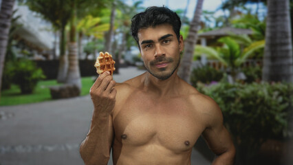 Young hispanic man sniffs golden waffle while holding it near bare chest in forest walkway;...