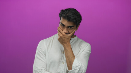 Fototapeta premium Young man in white shirt covers mouth with hand under purple studio lighting while tilting head downward; hesitation.