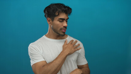 Young hispanic man adjusts white shirt with both hands over chest in blue studio setting;...