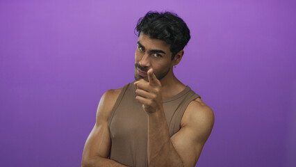 Naklejka premium Man pointing finger to eye in purple studio with bare muscular arms and sleeveless top showing toned biceps; insight clarity precision determination.