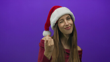Woman in red santa hat and burgundy off shoulder top beckoning with finger and winking in studio under purple light; playful invitation.