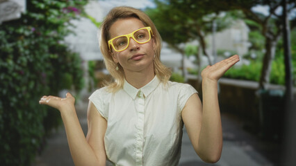 Woman with yellow glasses raises both hands palm up on sunlit park walkway under leafy green trees;...