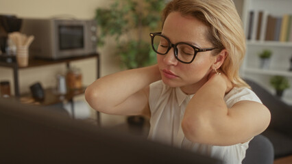 Blonde woman with glasses and eyes closed holds neck near computer in office building; stress...