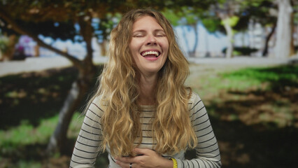 Obraz premium Happy woman covers hand while laughing in the park with blonde young hair wearing a striped watch outdoors.