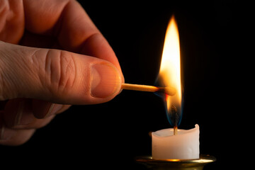 Close-up of hand lighting white candle with matchstick flame in dark background, capturing moment of ignition.
