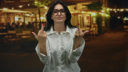 Young hispanic woman wearing white striped shirt and glasses raising both middle fingers while standing on street cafe terrace under string lights; anger.