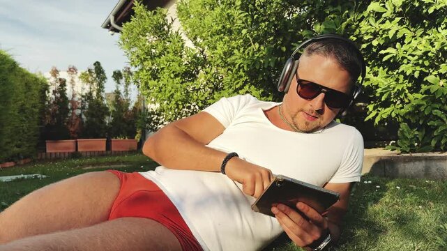 Man lying on lawn with tablet and headphones at sunset