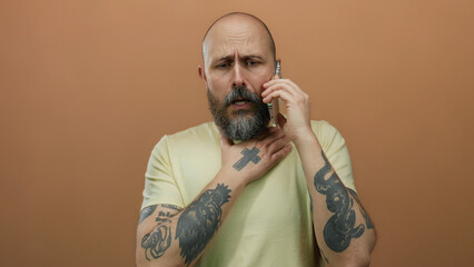 Man with beard and tattoos talks on phone, touching throat in discomfort against brown wall, wearing casual shirt.