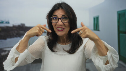 Woman finger pointing to self with smiling face on seaside street wearing white blouse and glasses;...