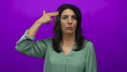Fototapeta premium Hispanic woman pointing finger gun to head in studio with purple background wearing green blouse and pearl necklace; frustration.