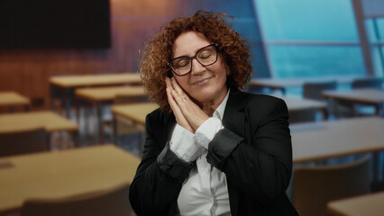 Woman stretching and gesturing sleepily in a classroom with wooden desks and modern design, wearing...