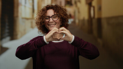 Woman with curly hair and glasses makes heart gesture while smiling on a city street background,...