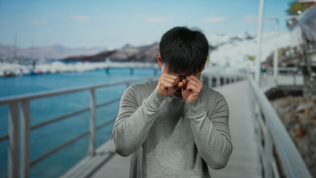 Young man on seaside promenade smiling and rubbing eyes, wearing casual clothing, with a scenic beach and clear blue sky in the background.