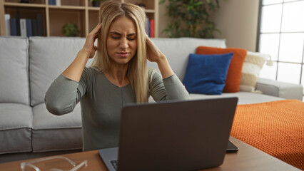 Young blonde woman holds temples and grimaces over laptop at home desk beside bookshelf in living room; stress pressure productivity.