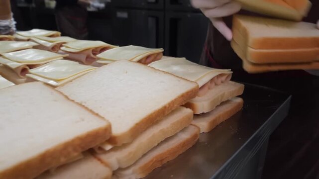 A person with gloved hands rapidly adds rolls of cold cut meat and slices of yellow cheese onto a large, organized grid of bread in a commercial kitchen environment.
