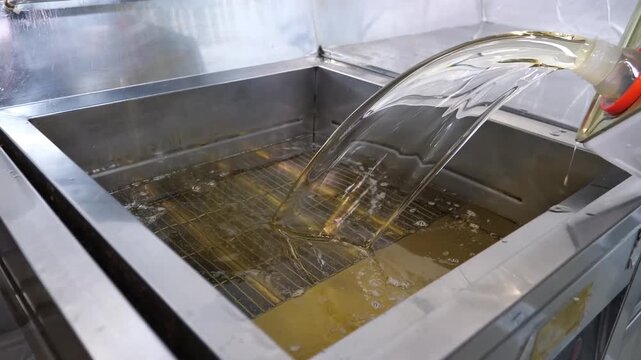 A chef in a commercial kitchen pours oil into a deep fryer, then lowers two baskets of fresh potato slices into the hot liquid to begin frying them until golden and crispy.