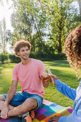 Friends raising beer bottles, sharing jovial moment during sunny park picnic, celebrating...