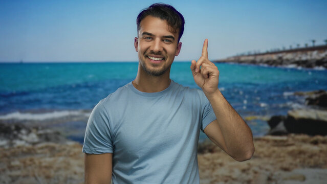 Young hispanic man smiling at a seaside beach, pointing confidently with the ocean and blue sky in the background, conveying a sense of joy and leisure outdoors.