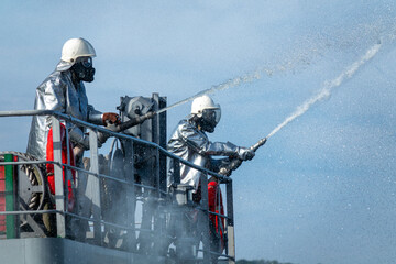 Two firefighters in silver heat-resistant suits and gas masks spray water from hoses during an...