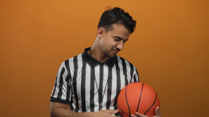 Young hispanic man wearing referee uniform holding basketball against orange background embodying athletic referee persona in isolated vibrant setting.