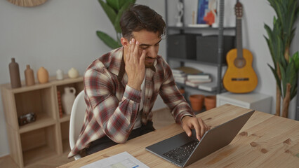 Hispanic man working on a laptop in a modern office, displaying stress amidst an interior with plants and a guitar in the background, reflecting a busy workplace environment.