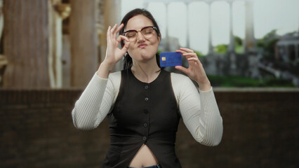 Woman making ok gesture while proudly showing credit card in front of ancient roman ruins under...