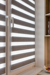 Daylight-filtering zebra roller blind in a kitchen interior. Sheer and solid fabric stripes of roller shades alternate to adjust brightness and privacy while complementing modern decor and countertop.