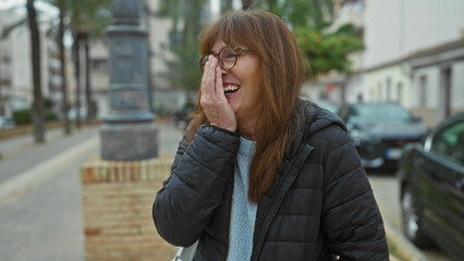 Fototapeta premium Woman laughing joyfully covering face in city street with cars and trees, wearing glasses and jacket in an outdoor setting on a chilly day.