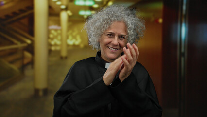 Woman clapping in a hotel lobby, expressing joy, wearing clerical attire, with a vibrant smile,...